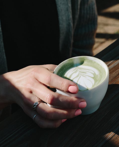 A hand holding a matcha tea latte with steamed milk flower art in a coffee mug
