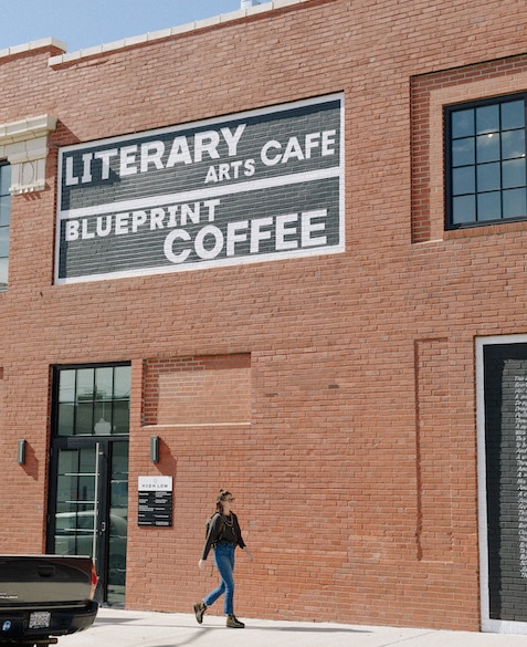 Nora Brady walks on the sidewalk next to Blueprint Coffee High Low in front of the sign that reads "Literary Cafe"