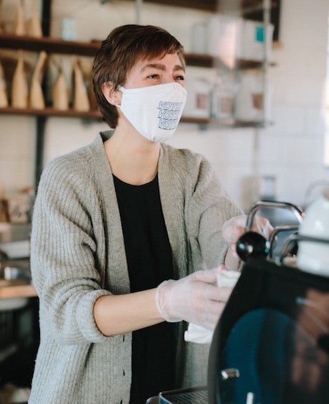 MG 2 MG Batts smiles under a white mask with "vote" on it in blue while brewing coffee