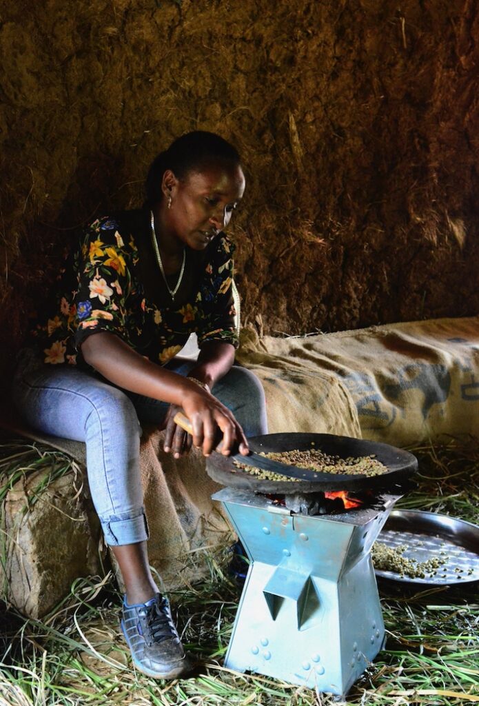 A woman sitting down roasts coffee beans over a fire.