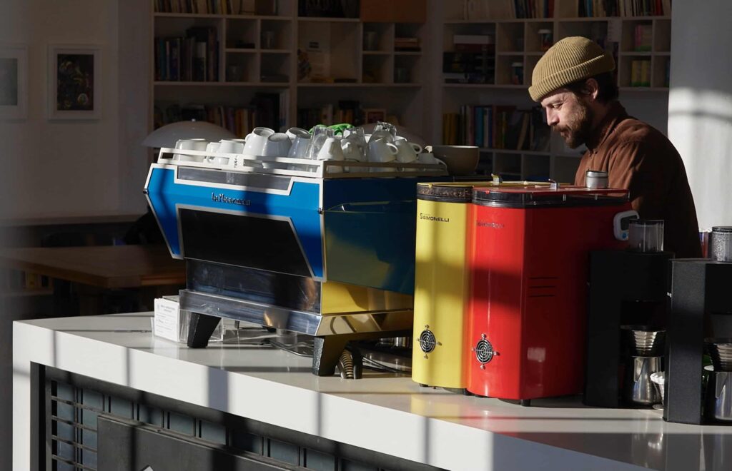 A smiling barista works behind a counter. On the counter sits a blue espresso machine and red and yellow coffee grinders.