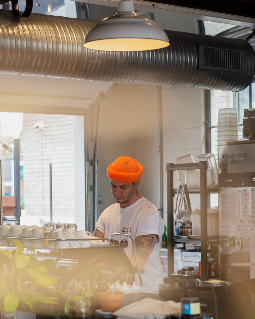 Britton wears an orange hat and looks down at the espresso machine at Watson
