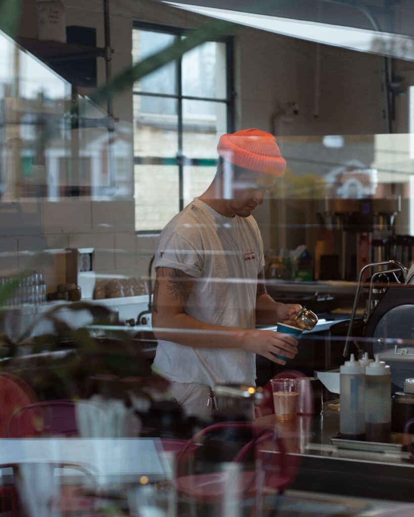 Britton steaming milk at the espresso machine at Watson, viewed from outside looking in through the window