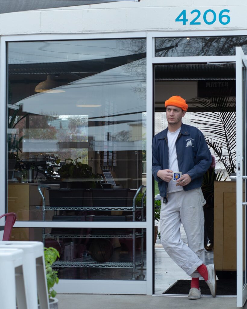 Britton leans against the door frame of the open back door at Watson, holding a coffee mug