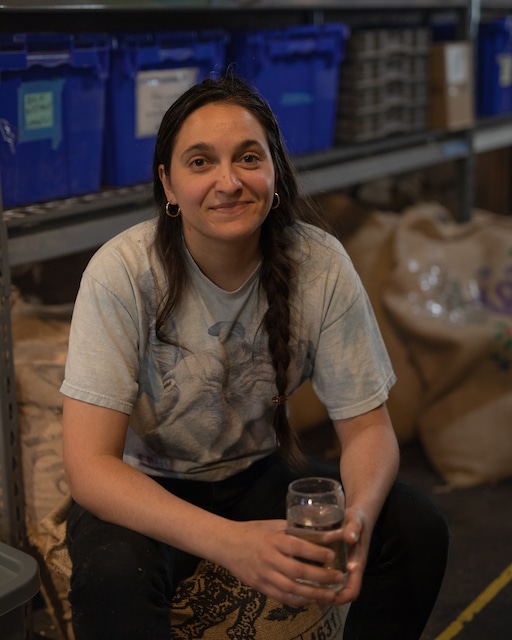 Marija sits in front of a shelf of burlap sacks of green coffee, looking at the camera and smiling