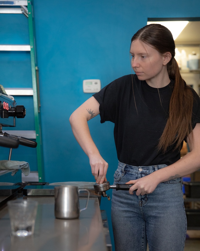 Val behind the bar at our Delmar café, tamping espresso in a portafilter resting on the edge of the countertop