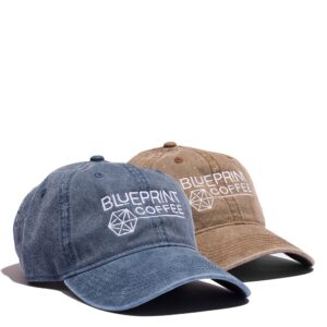 Two hats against a white background. A blue version of the Blueprint "Dad" hat on the left with the Blueprint Coffee logo in white. The brown hat on the right features the same logo.