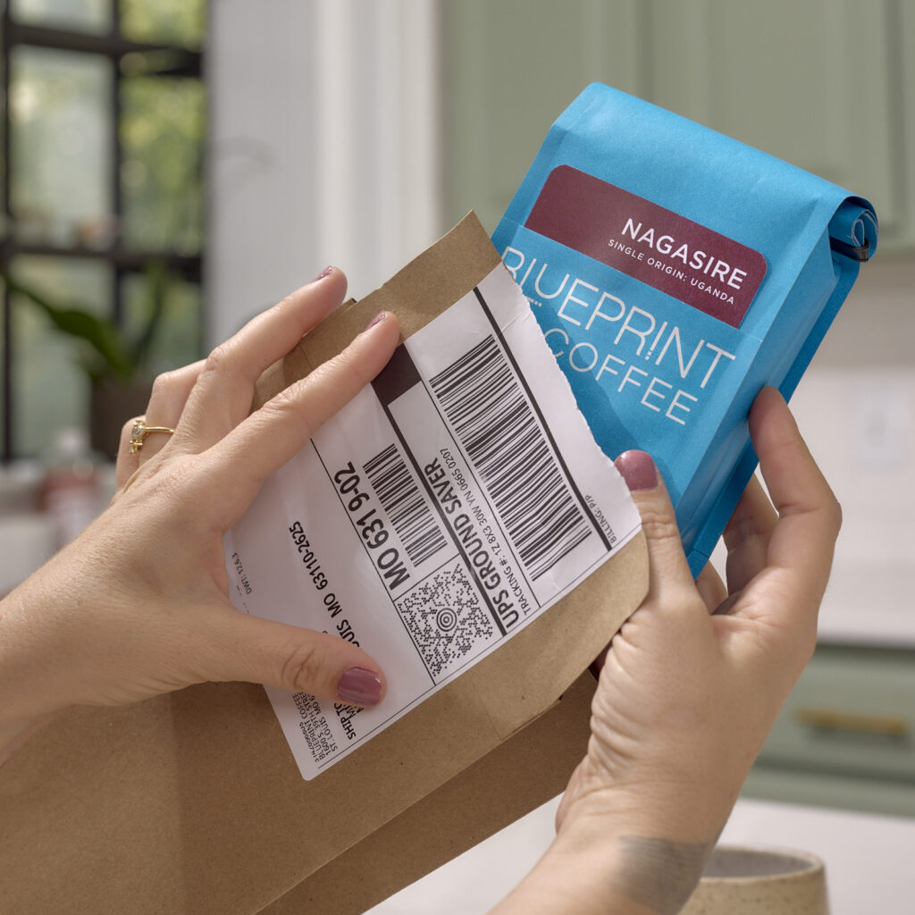 A person removes a blue 10 oz bag of Blueprint Coffee beans from their subscription mailing envelope