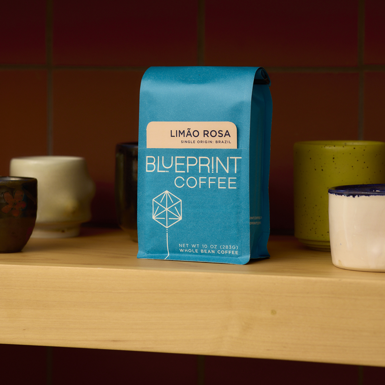 A bag of Limão Rosa, Brazil, roasted by Blueprint Coffee, sits on a wooden shelf among handmade ceramic mugs.