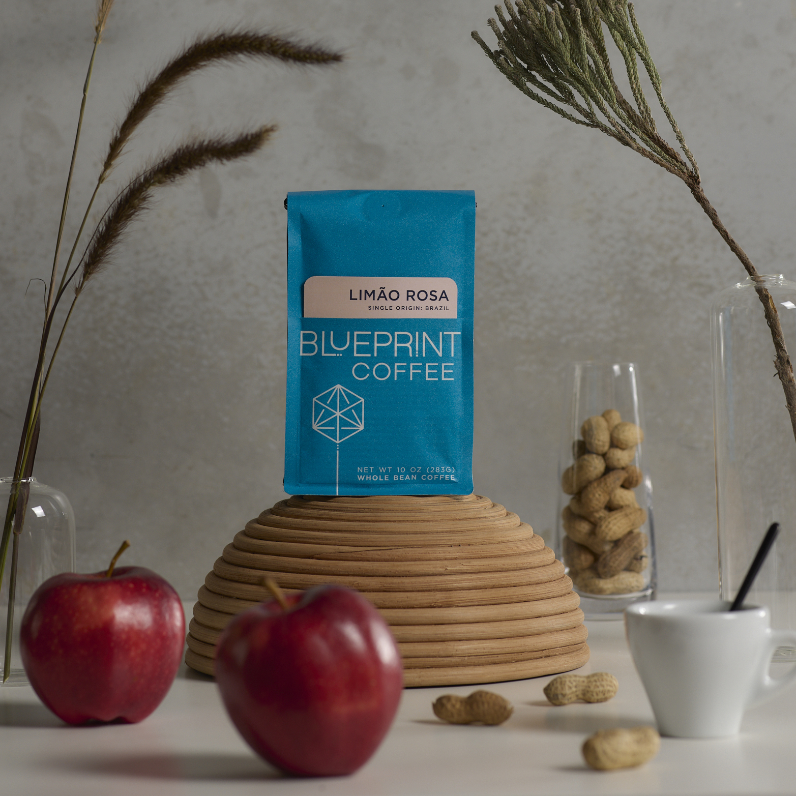 A bag of Limão Rosa, Brazil, roasted by Blueprint Coffee, sits atop an upturned bowl, surrounded by apples, peanuts, and dried flowers arranged in a still life composition.