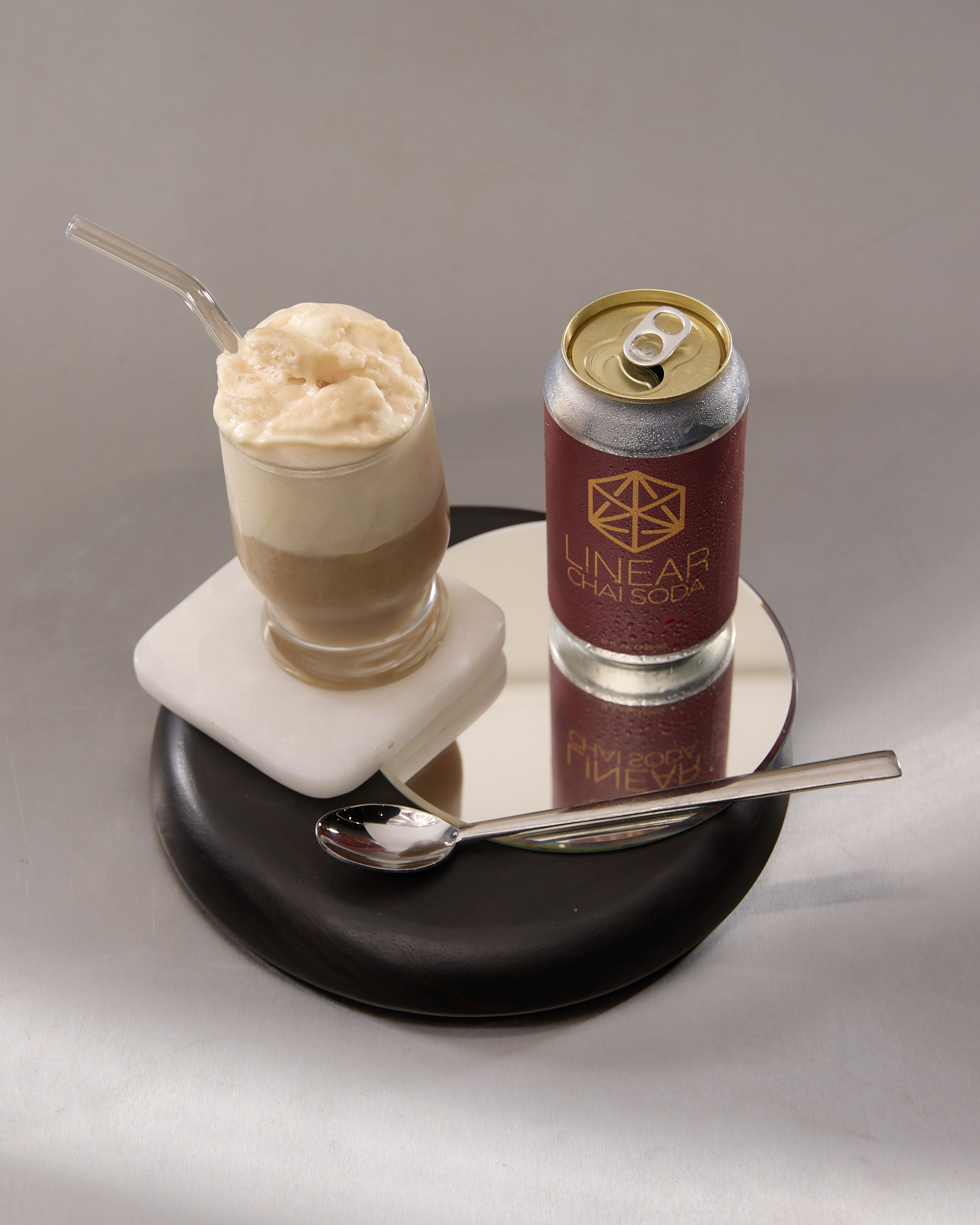 A Chai Soda float, made from Linear Chai Soda and vanilla ice cream, in a glass and arranged on a tray beside its empty dark red can and a metal spoon