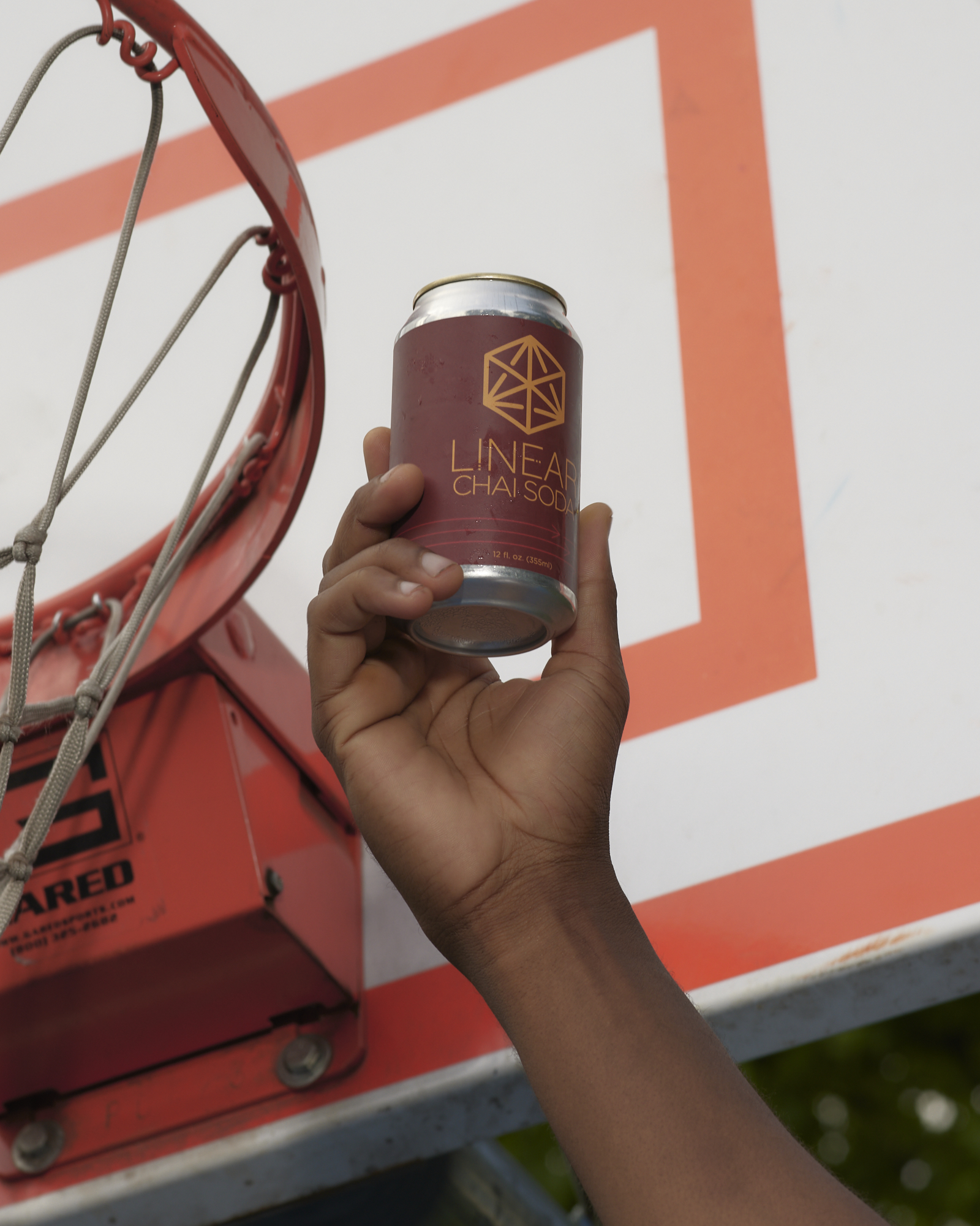 A person's hand holds up a dark red can of Linear Chai Soda beside the orange rim and white backboard of a basketball goal