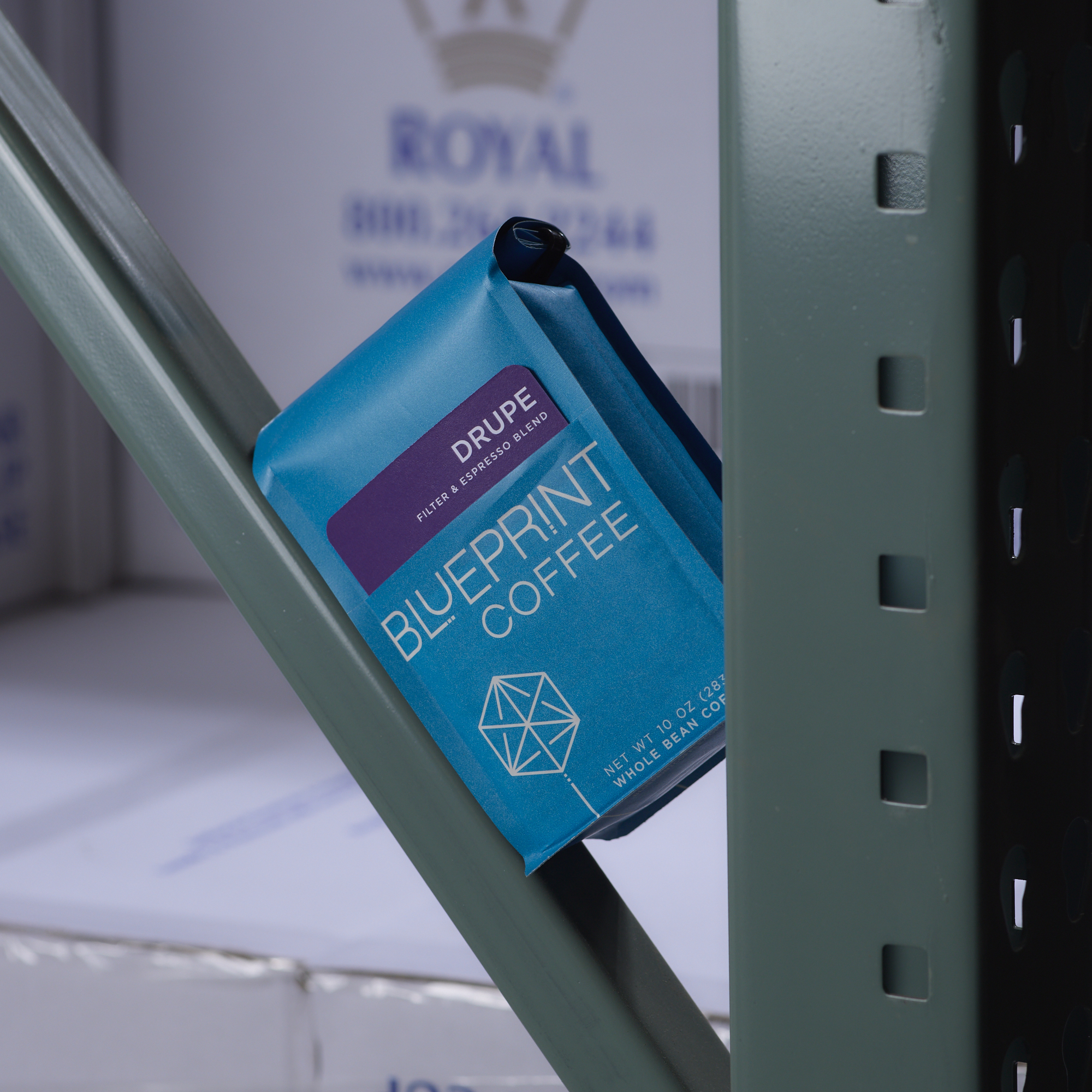 A bright blue 10 oz bags of Drupe Filter & Espresso Blend sits wedged in the green metal supports of a warehouse shelf