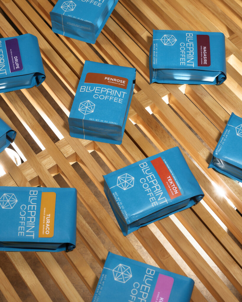 Bright blue bags of Blueprint Coffee beans are arranged on a surface of wooden slats. Pink, yellow, orange, red, and brown cards are tucked into a pocket on the front of each bag, labeling the coffees inside.