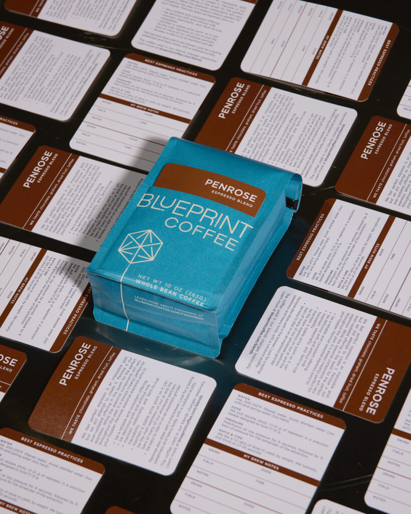 A bright blue bag of Penrose coffee beans from Blueprint Coffee is in the center of the frame, surrounded by the cards that are used to label the bag, arranged to show both the front and back of the cards.