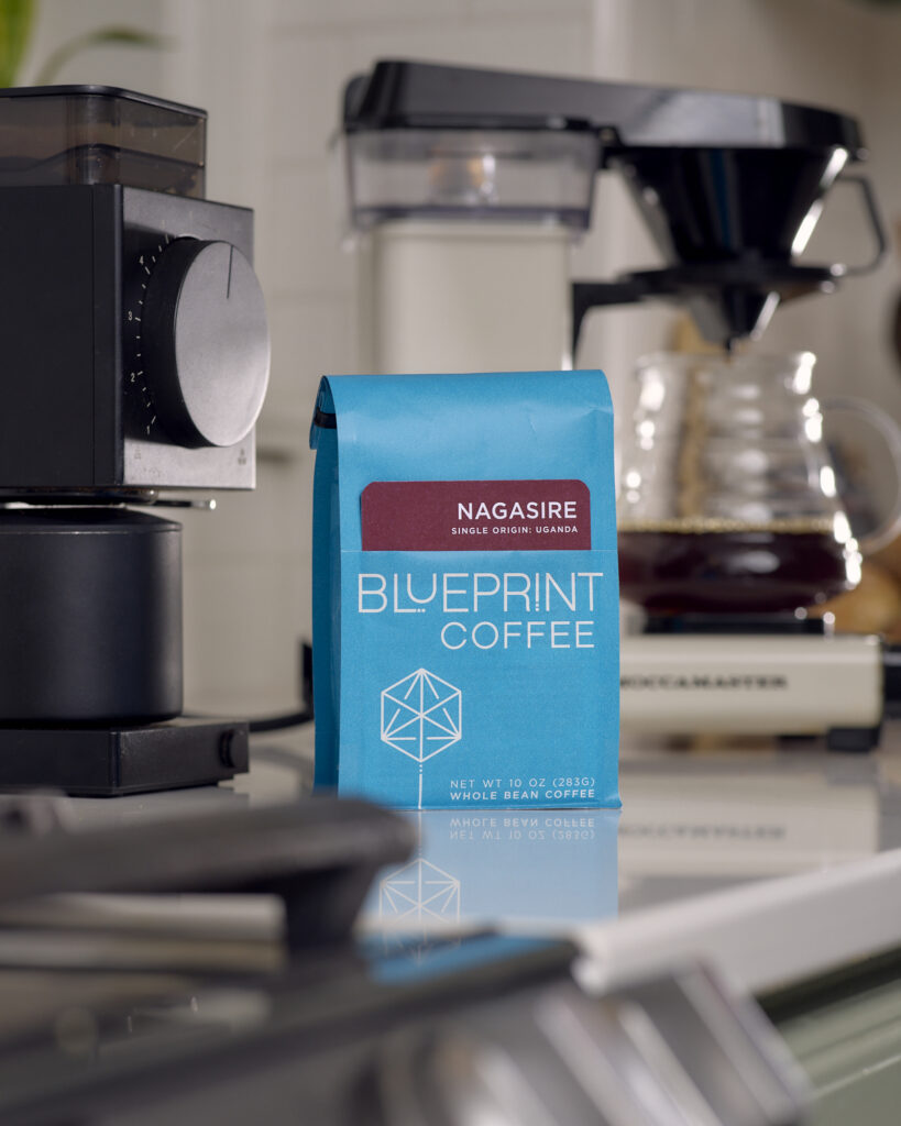 A bright blue bag of Nagasire, Uganda coffee beans from Blueprint Coffee in the foreground on a home kitchen counter. A freshly brewed cup of the coffee is behind it.