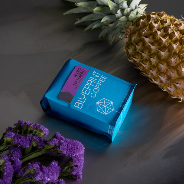 A bag of Raro Nansebo instant coffee sits on a table with lavender flowers and a pinapple.