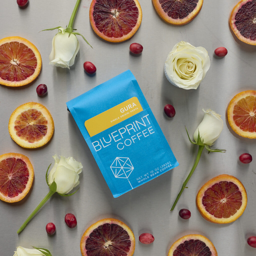 A blue 10-ounce bag Gura, Kenya laid flat with an arrangement of blood orange slices, cranberries, and white roses surrounding it