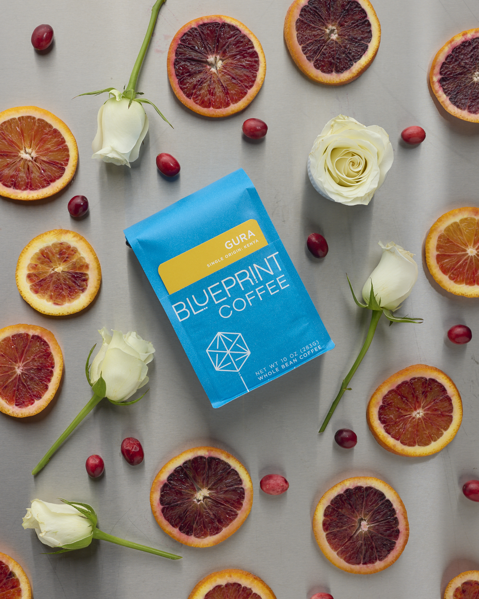 A blue 10-ounce bag Gura, Kenya laid flat with an arrangement of blood orange slices, cranberries, and white roses surrounding it