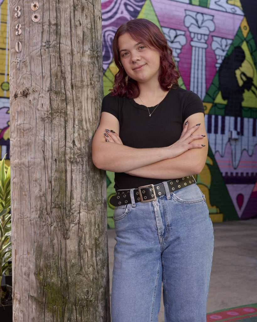 Sylvia stands with her arms crossed, leaning against a wooden light pole