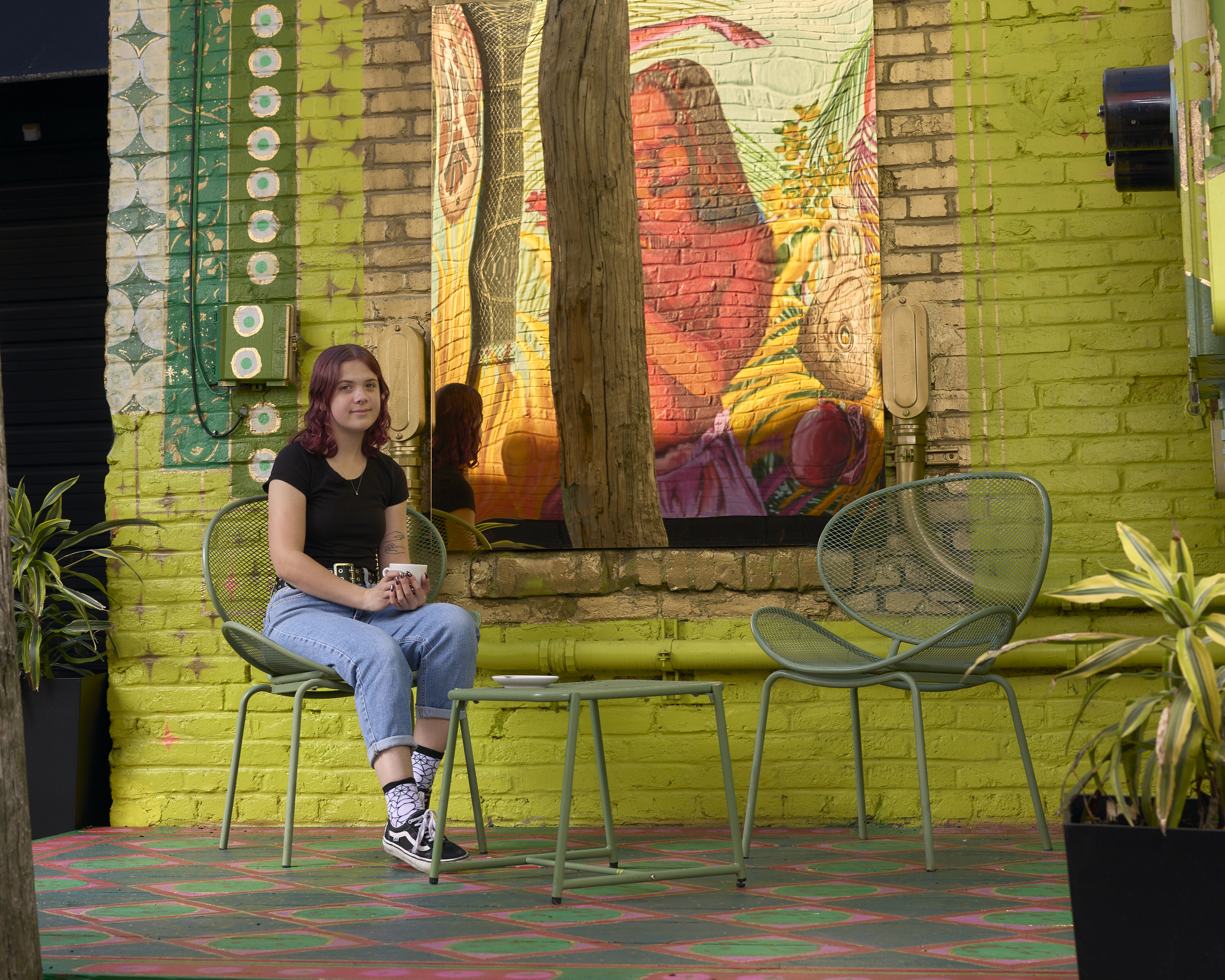 Sylvia sits at a table outside in front of a green wall painted with a large mural