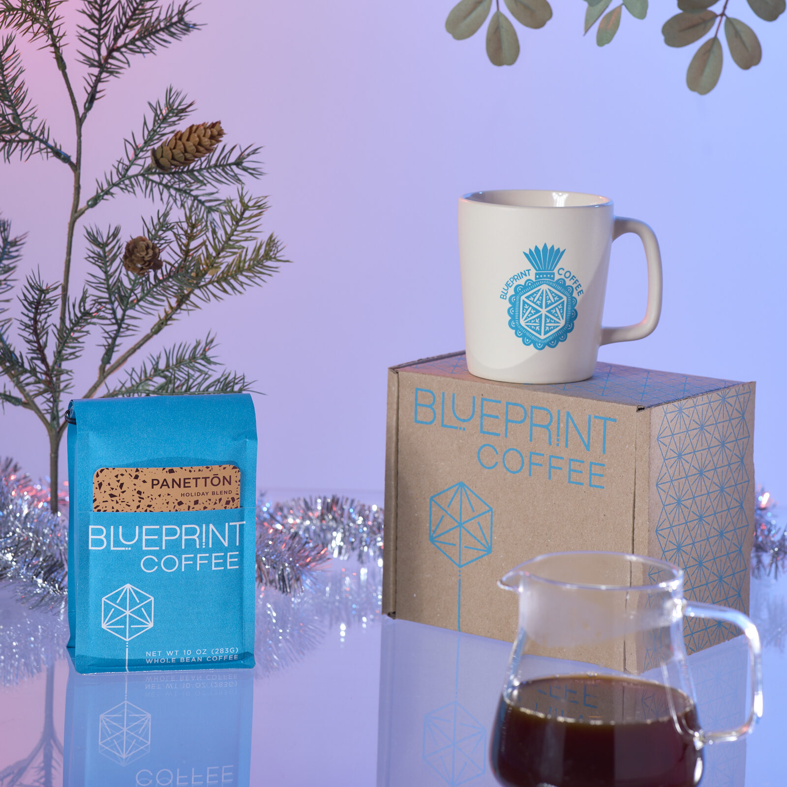An arrangement includes a screenprinted box with the Blueprint logo, a white mug with a blue screenprinted design, and a blue bag of Panettōn Holiday Blend coffee.
