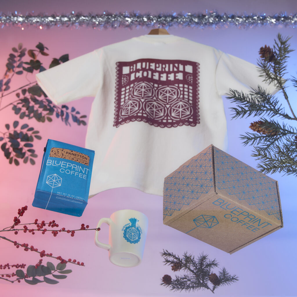An arrangement showing a screenprinted box with the Blueprint logo, a mug with a blue screenprinted design, a white cropped t-shirt with a red screenprinted design, and a blue bag of Panettōn Holiday Blend coffee.