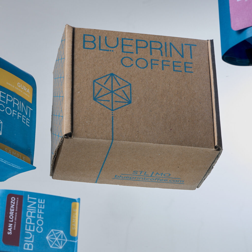 A brown box printed with the Blueprint Coffee logo is centered in the frame, with blue sample bags of single origin coffees arranged around it