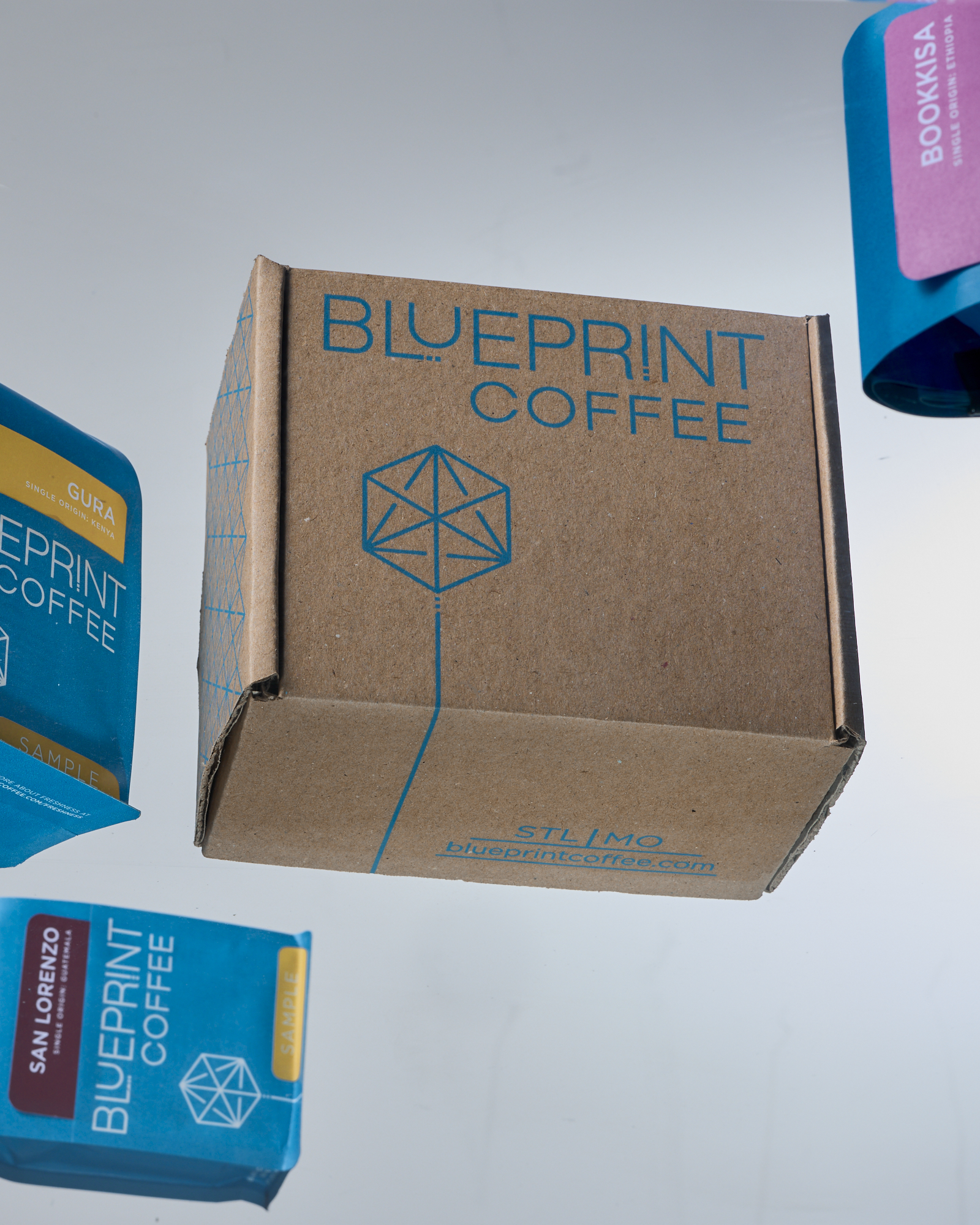 A brown box printed with the Blueprint Coffee logo is centered in the frame, with blue sample bags of single origin coffees arranged around it