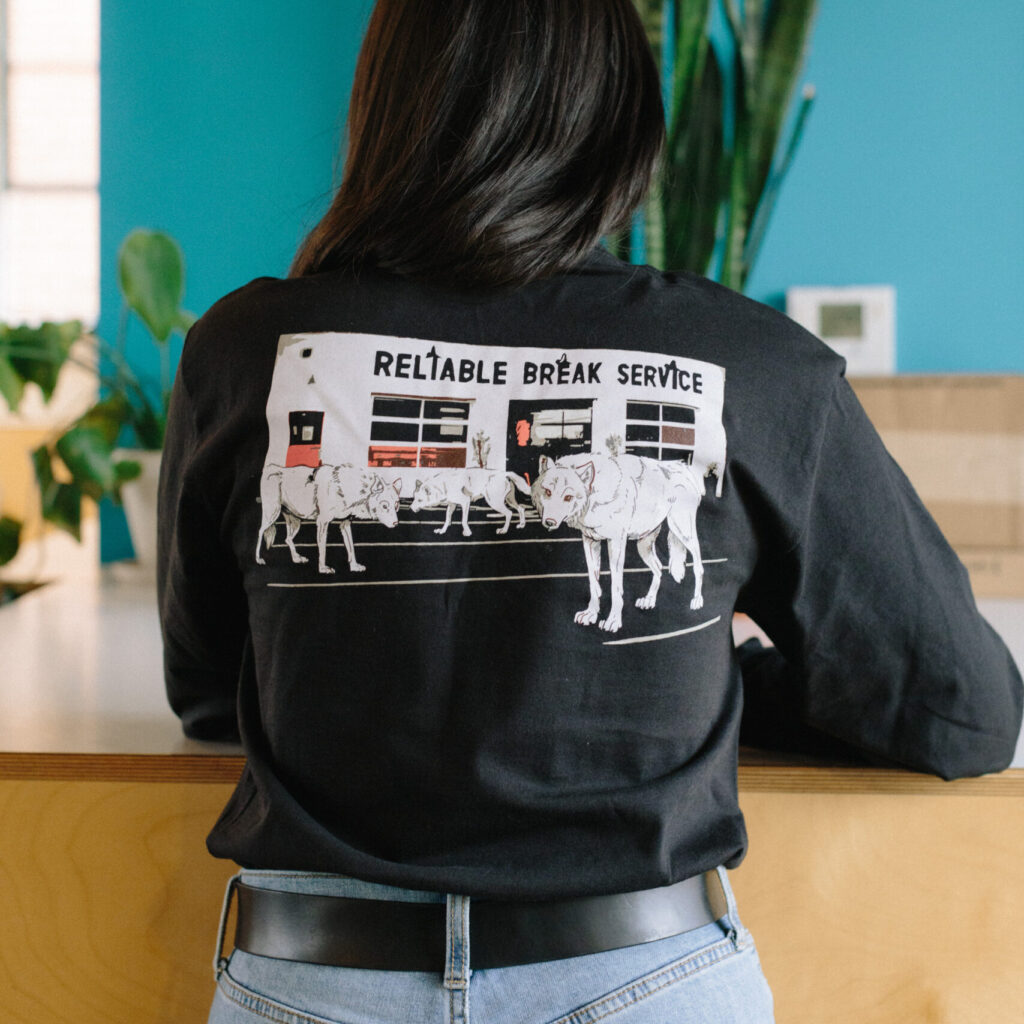 A person's back is turned to the camera, showing the illustration of wolves in front of our Watson café, printed in white on a black, long-sleeved t-shirt