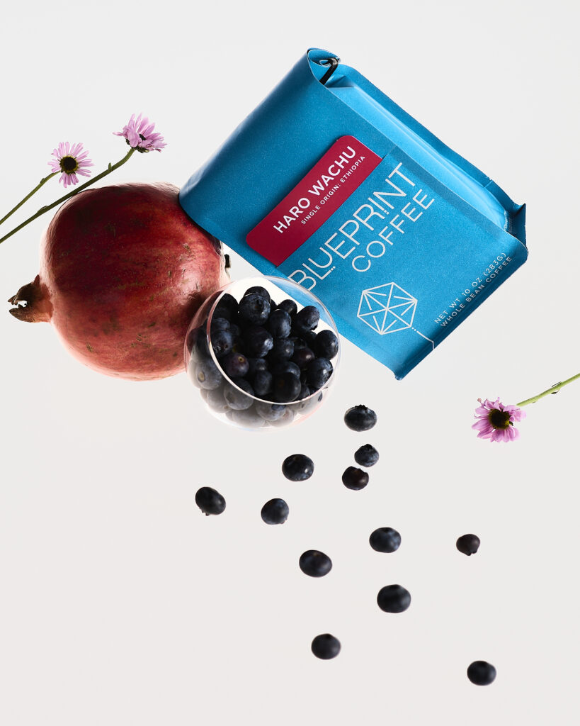 A bright blue bag of Blueprint Coffee beans with a removable red info card reading Haro Wachu, Ethiopia floats among an arrangement of blueberries, pomegranate, and pink flowers