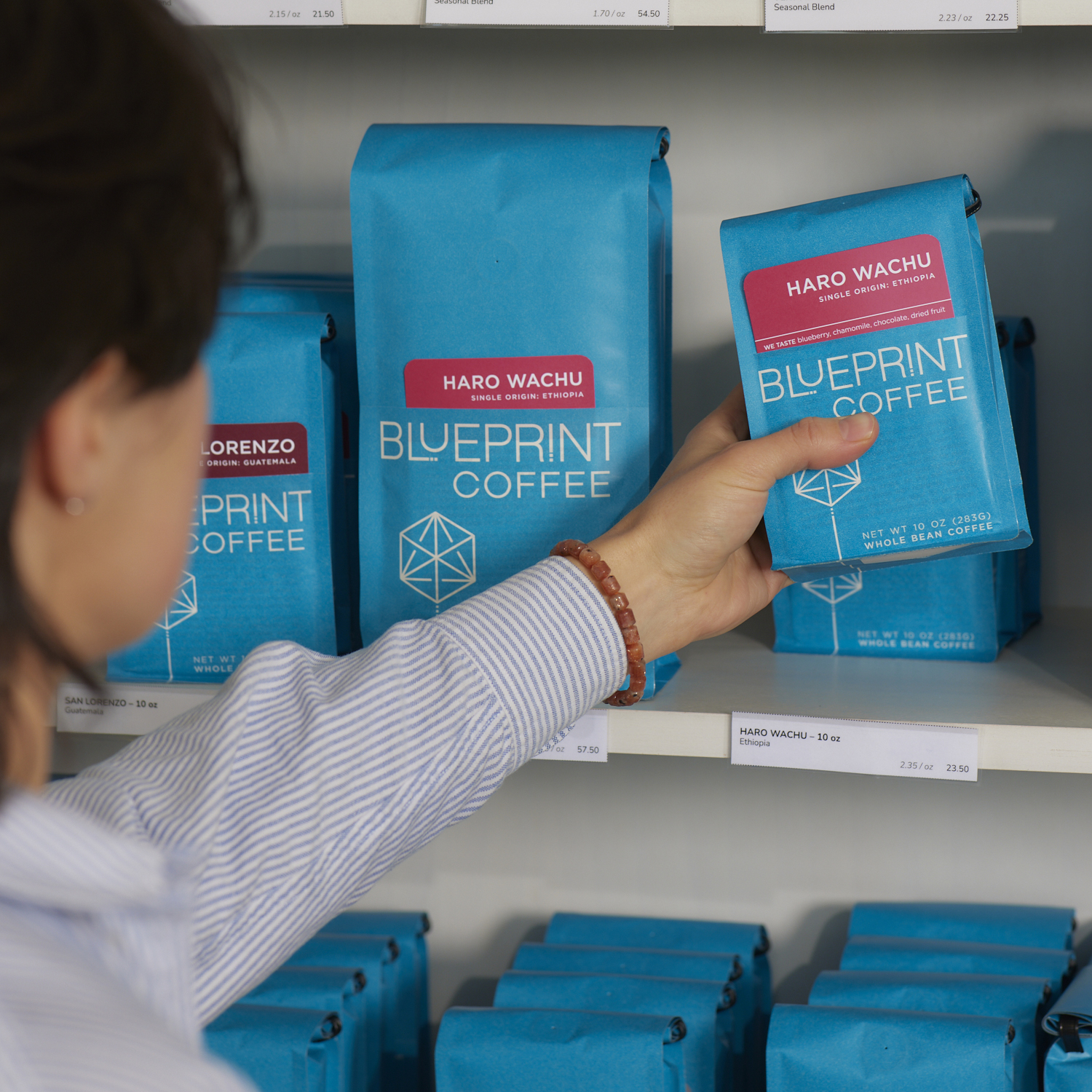 A person stands in front of a retail shelf stocked with coffees and teas. The person is grabbing a blue bag of Haro Wachu, Ethiopia coffee beans.