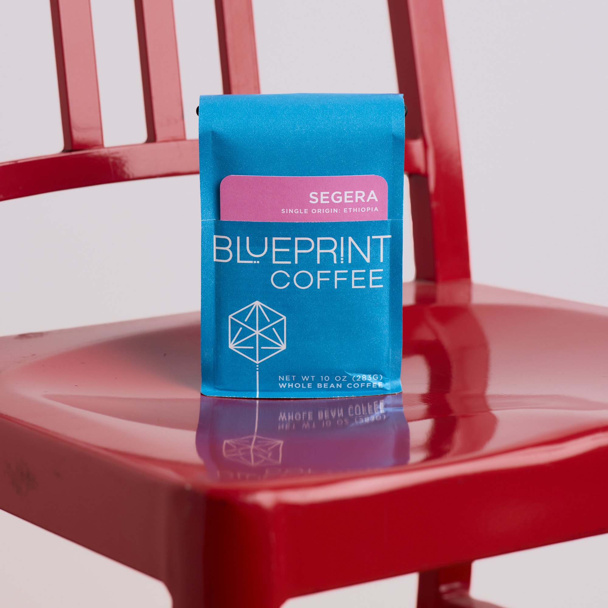 A bright blue bag of Segera, Ethiopia coffee beans with the Blueprint Coffee logo and a pink removable info card. The bag is placed on the seat of a bright red chair.