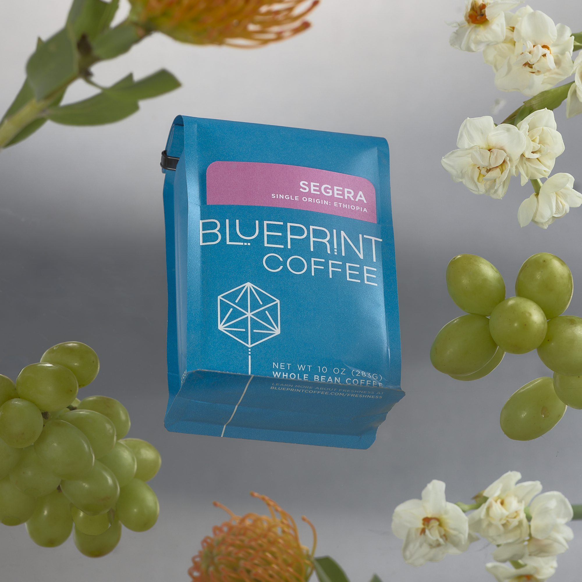 A bright blue bag of Segera, Ethiopia coffee beans with the Blueprint Coffee logo and a pink removable info card. The bag appears to be floating, with white flowers and green grapes surrounding it.