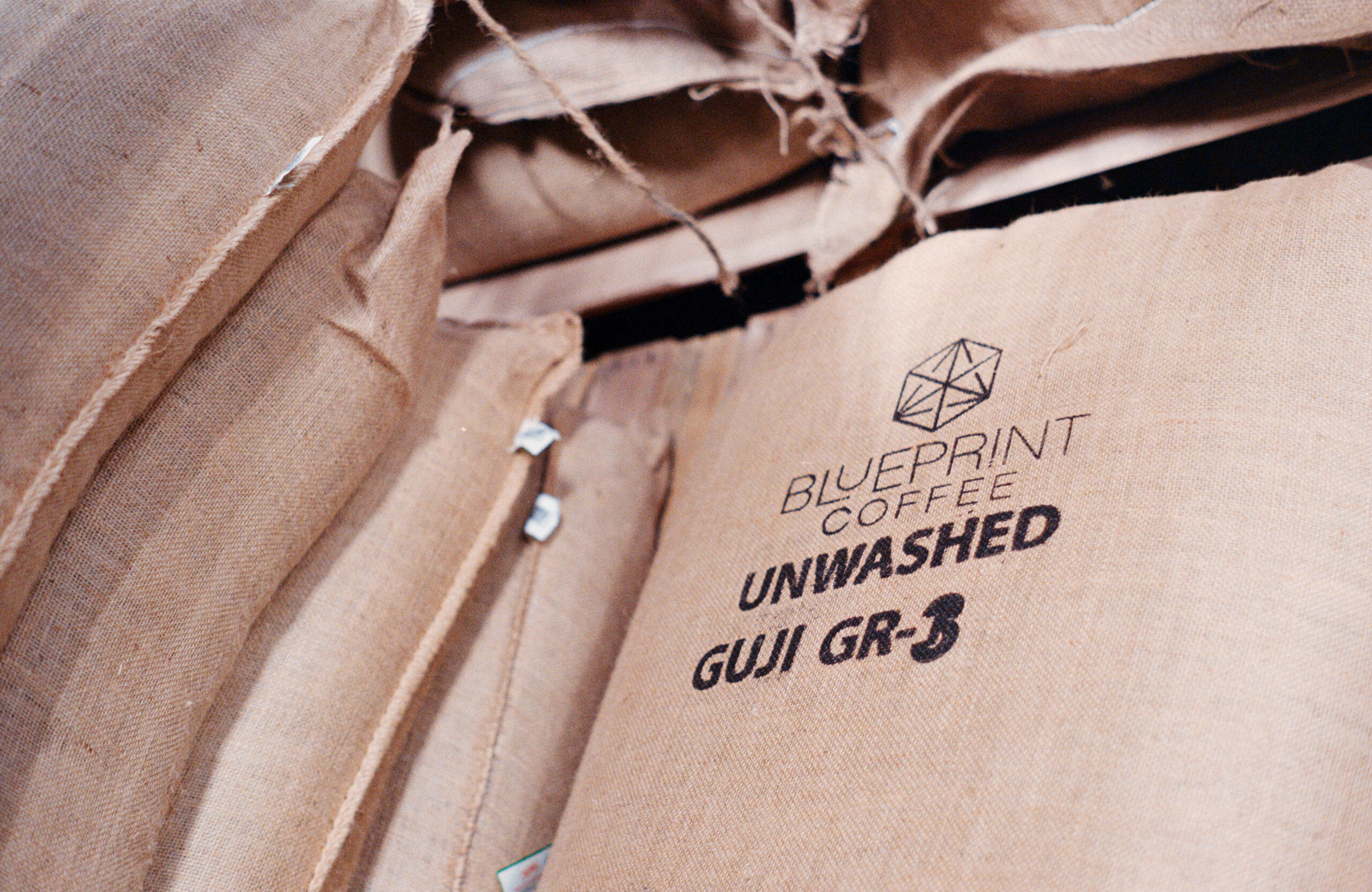 Burlap bags of green coffee are stacked flat on a warehouse shelf. The bag on top is printed with black text reading "UNWASHED GUJI GR-3" below the Blueprint Coffee logo.